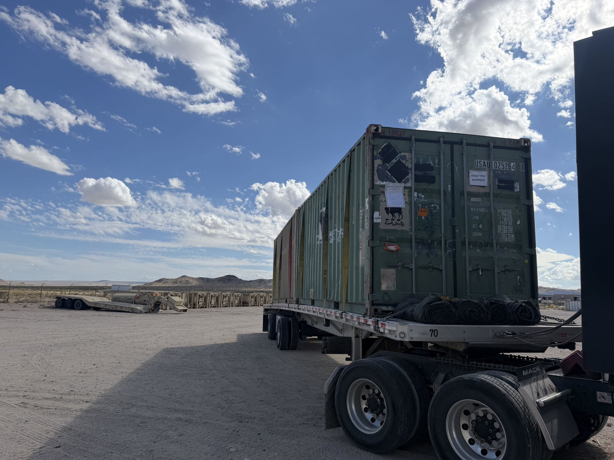 Flatbed trailer carrying a green shipping container in a desert yard with mountains in the background