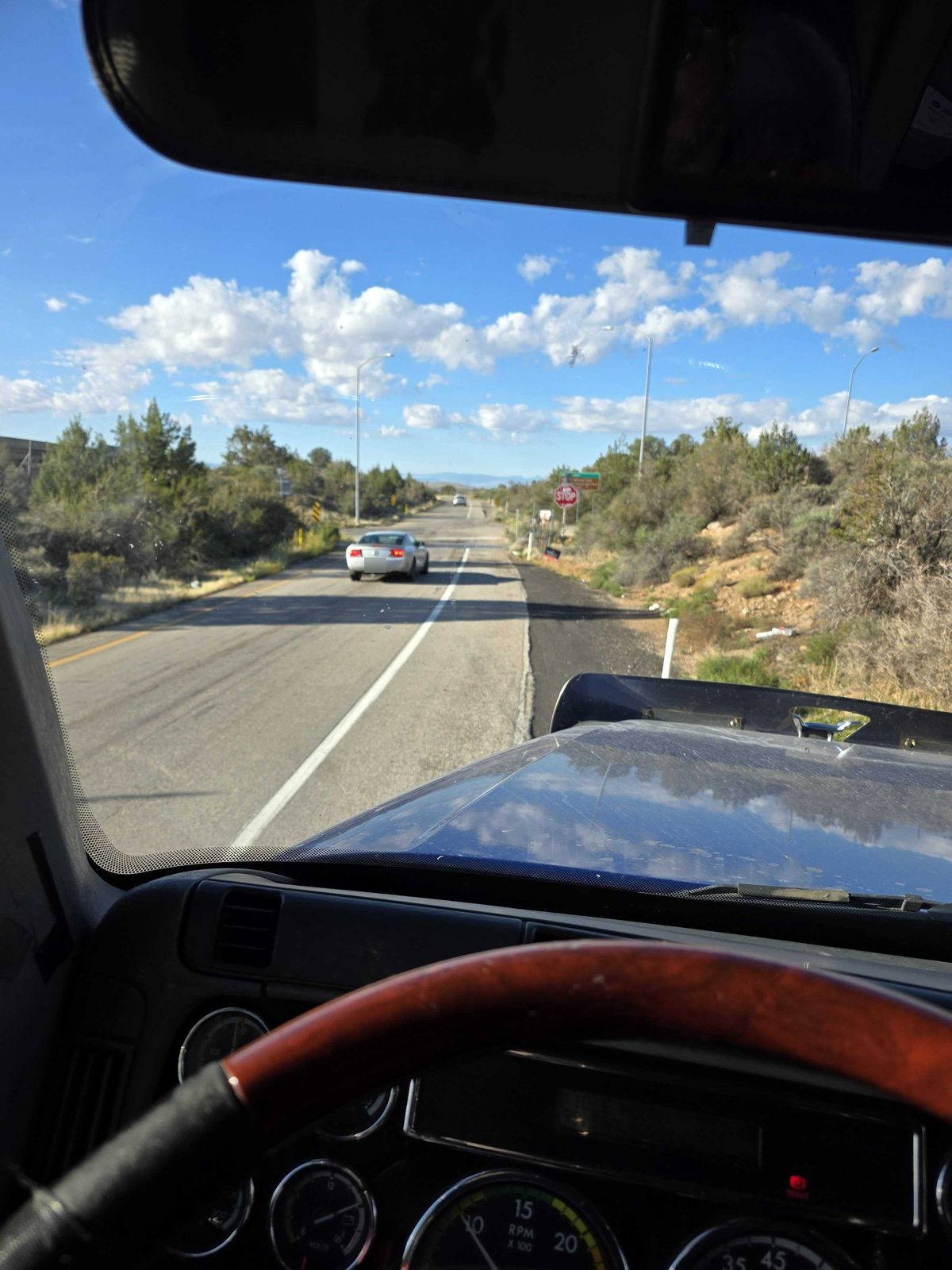 View through a truck windshield of a two-lane highway through high-desert scrub under a blue sky with white clouds