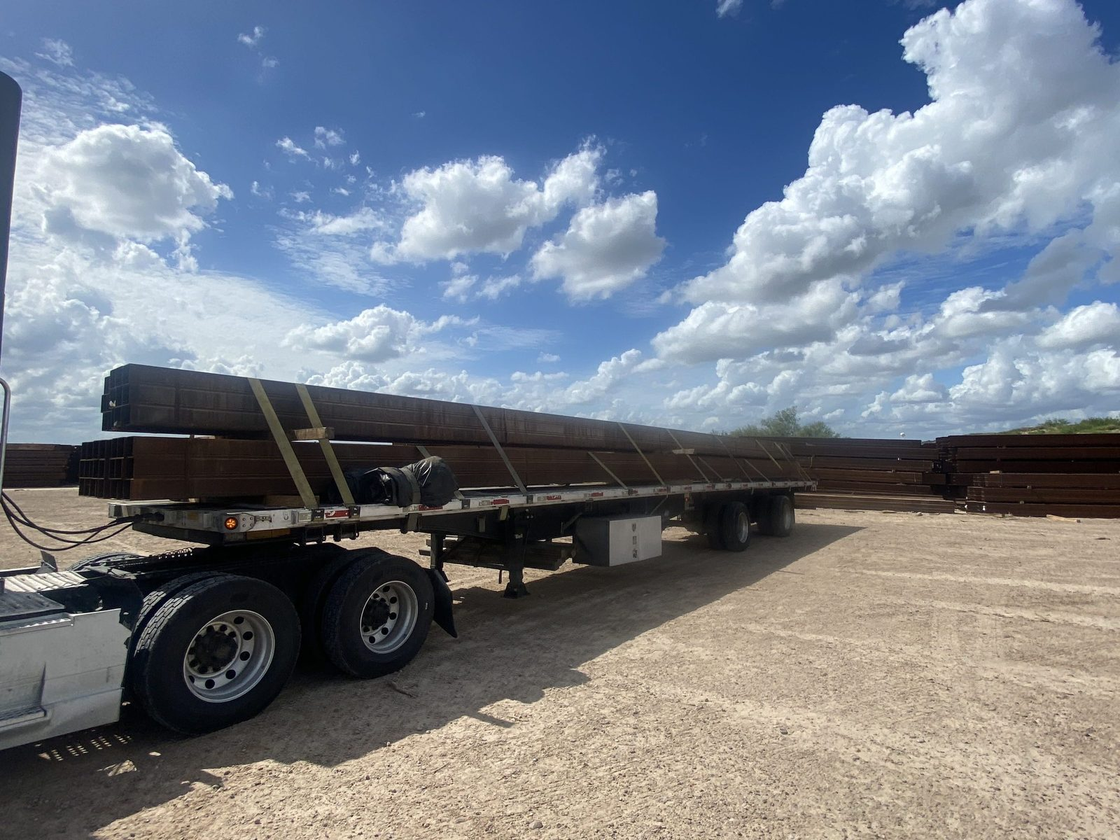 Flatbed trailer loaded with long steel beams in a dirt yard under a cloudy blue sky