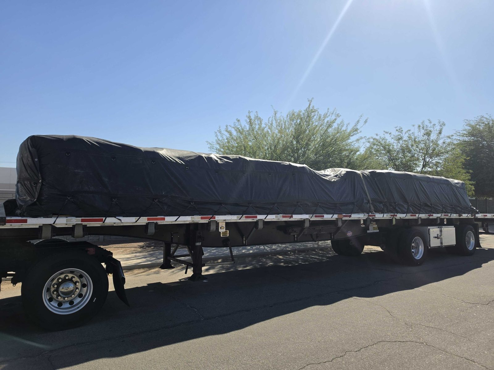 Flatbed trailer with a black-tarped load parked on a sunny street lined with desert trees