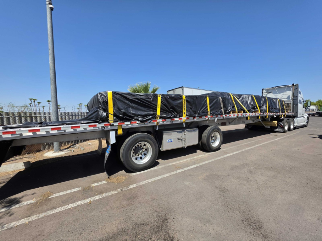 Side view of a flatbed trailer and semi truck showing trailer tires under a tarped, strapped load