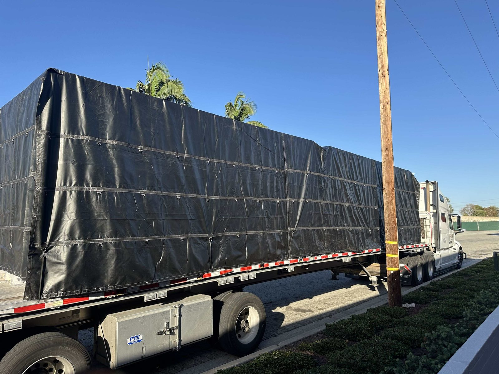 Full rig with a black-tarped flatbed load parked curbside under palm trees