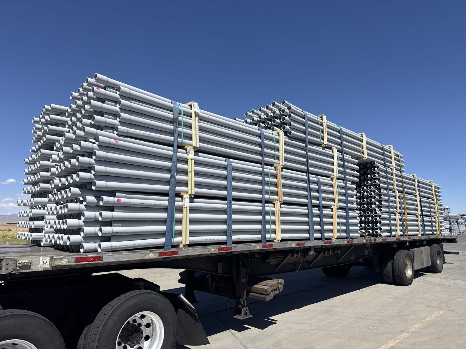 Flatbed trailer loaded with strapped bundles of gray pipe under a clear blue sky