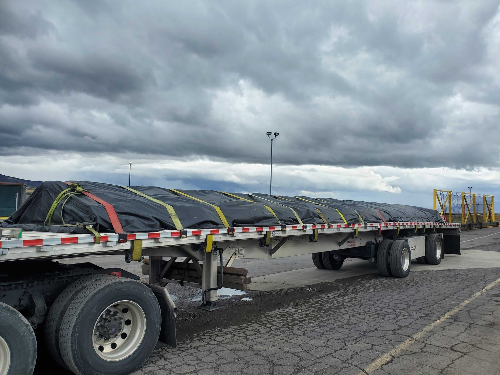 Flatbed trailer with a black-tarped, strapped load parked under storm clouds before an I-10 run
