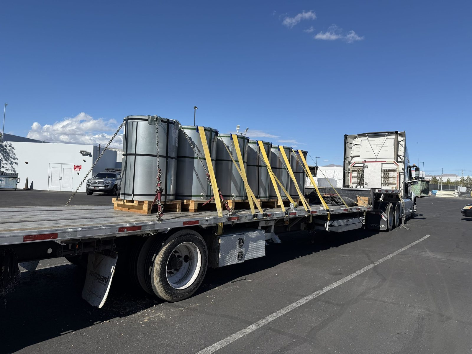 Flatbed trailer hauling steel coils on pallets secured with chains and yellow straps