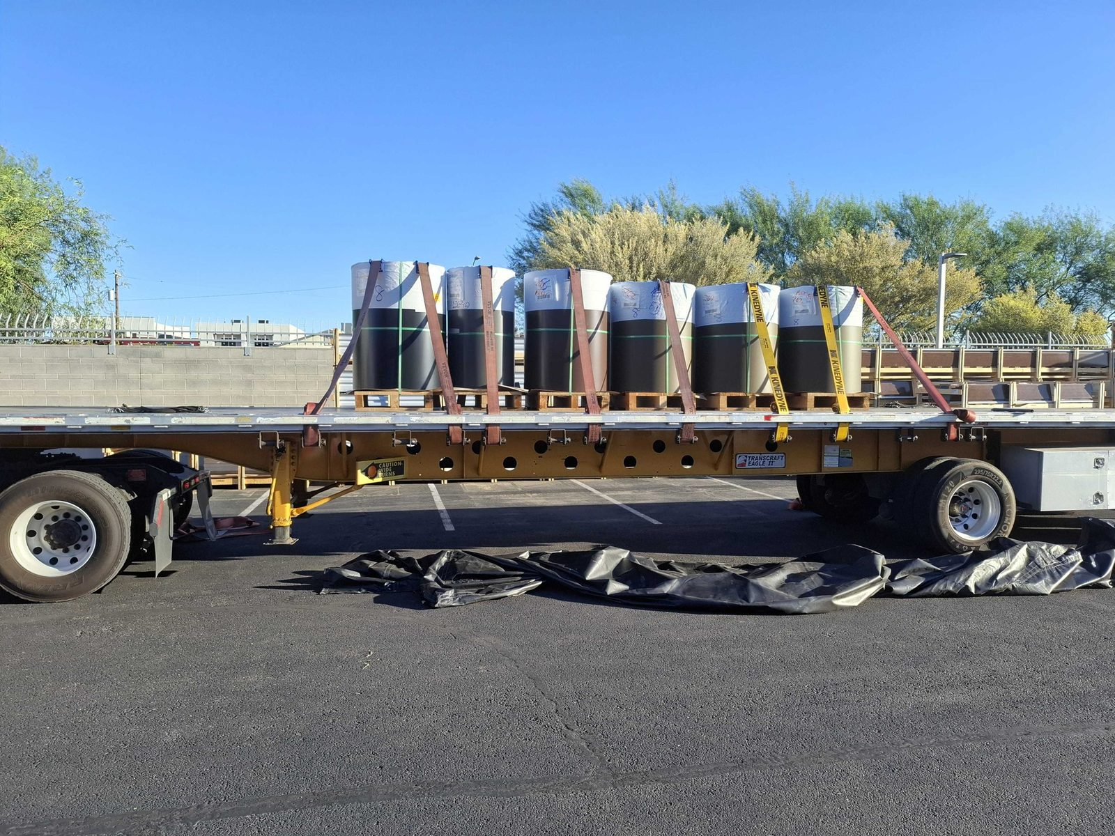 Flatbed trailer with a row of strapped drums parked under palm trees