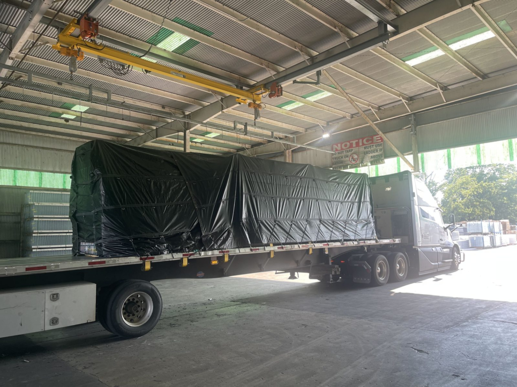 White semi truck and tarped flatbed load under a warehouse shed