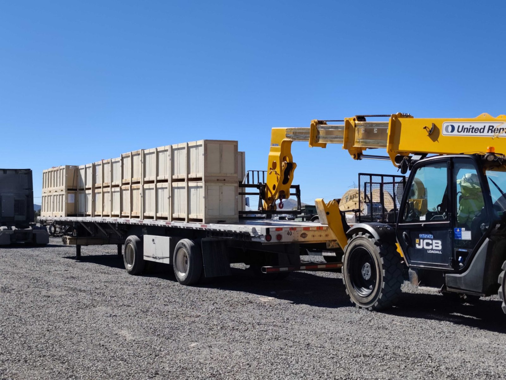 Telehandler unloading wooden crates from a flatbed trailer on a gravel lot